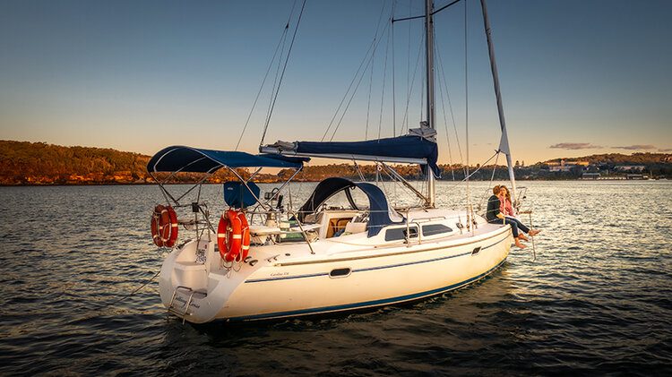 A sailboat with blue and white sails is anchored on a calm body of water during sunset. Two people are sitting at the back, enjoying the scenic view. Life preservers are visible on the boat, and an enchanting shoreline with trees is seen in the background.