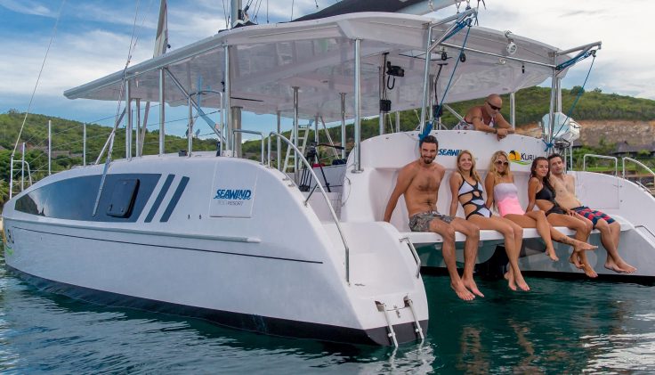 Five people in swimsuits sit on the edge of a white catamaran boat named "Seawind," with their feet in the water. The boat is docked in a tranquil, scenic area with lush greenery and cliffs in the background. Nearby, a man stands on deck gazing at an enchanting sunset over the water.