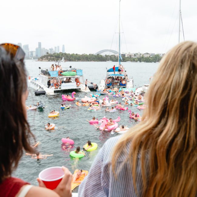 People on a boat enjoying a lively scene with numerous individuals floating on inflatable rafts in the water. The background features additional boats and a distant city skyline under a cloudy sky. Experience this vibrant atmosphere with Luxury Yacht Rentals Sydney from The Yacht Social Club.