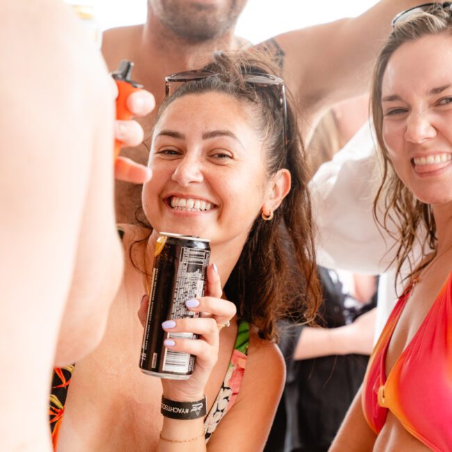 Two women in swimwear smile and pose for the camera at a lively gathering by The Yacht Social Club Event Boat Charters. The woman on the left holds a can of drink, and both appear to be enjoying a sunny day. There are other people around, suggesting a social event or party atmosphere.