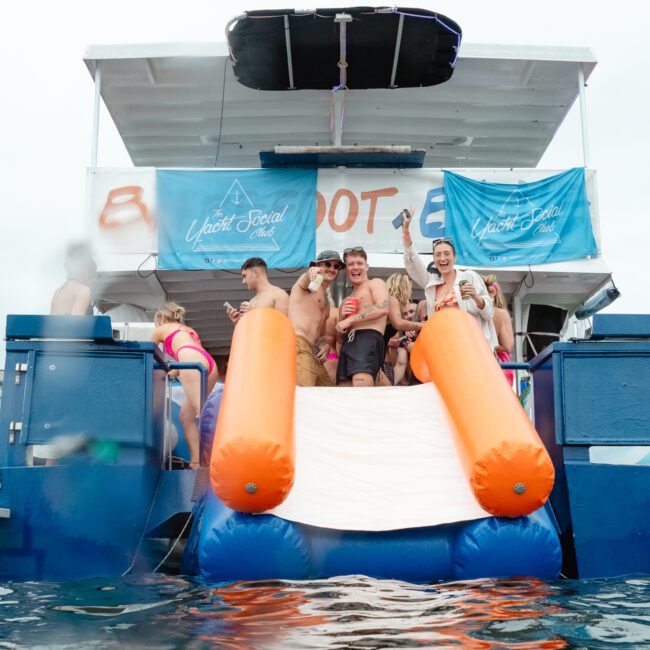 A group of people enjoys a party on a docked boat. Several individuals are standing and holding drinks, smiling at the camera. A large inflatable slide is attached to the back of the boat, leading into the water. Two blue banners from The Yacht Social Club Sydney Boat Hire hang on the boat.