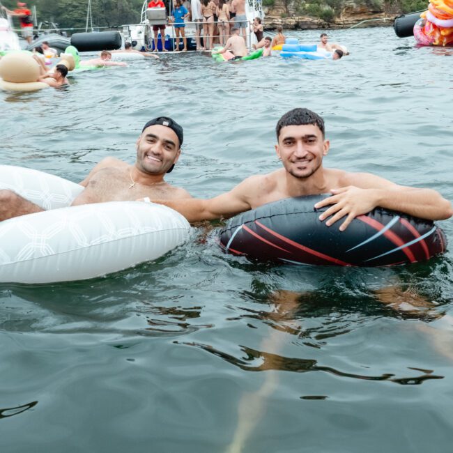 Two men are in an outdoor body of water, each sitting in an inflatable ring. They are smiling at the camera and holding onto each other's rings. In the background, more people float on inflatables near boats and a rocky shore, enjoying a day out with The Yacht Social Club Event Boat Charters.