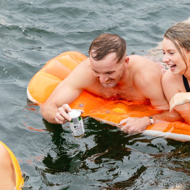 Two adults, a man and a woman, are having fun on an orange inflatable float in the water, holding drinks and smiling. Another person, partially visible, is swimming nearby, adding to the cheerful scene – it’s part of The Yacht Social Club Event Boat Charters experience.