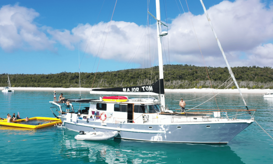 A sailing boat named "Major Tom" is anchored in clear, turquoise waters near a sandy beach. Several people are on the boat, with others enjoying themselves on a vibrant yellow inflatable raft and a small white dinghy. Lush green trees stand tall in the background under a partly cloudy sky.