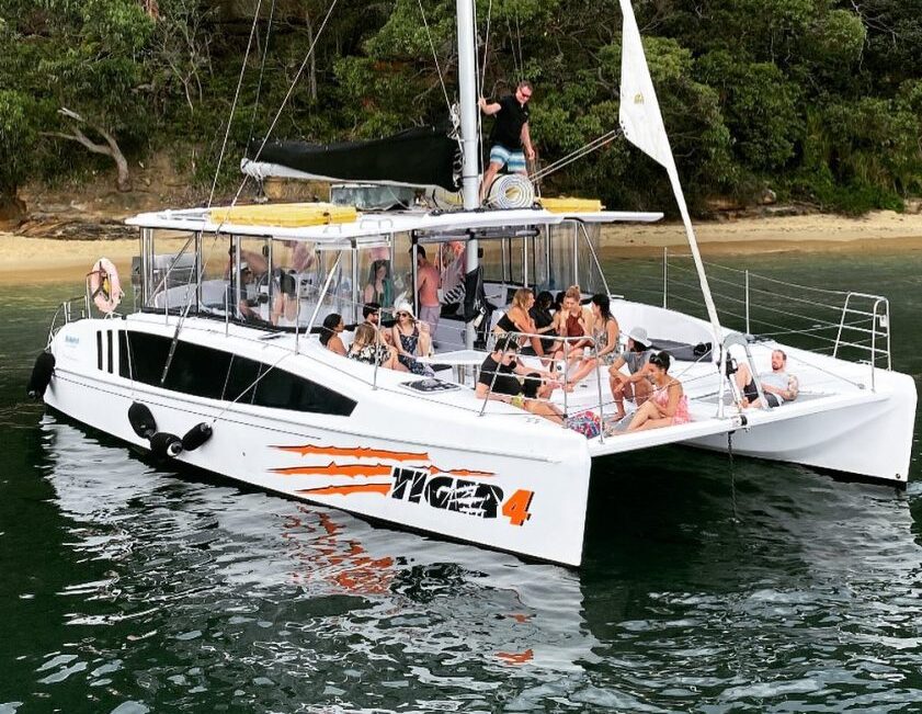 A large catamaran named "Tigger 4" is anchored close to the shore with lush green trees in the background. Several people are on board, enjoying the day. Some sit on the deck while others are standing or lounging. The calm water around the boat reflects its vibrant atmosphere.