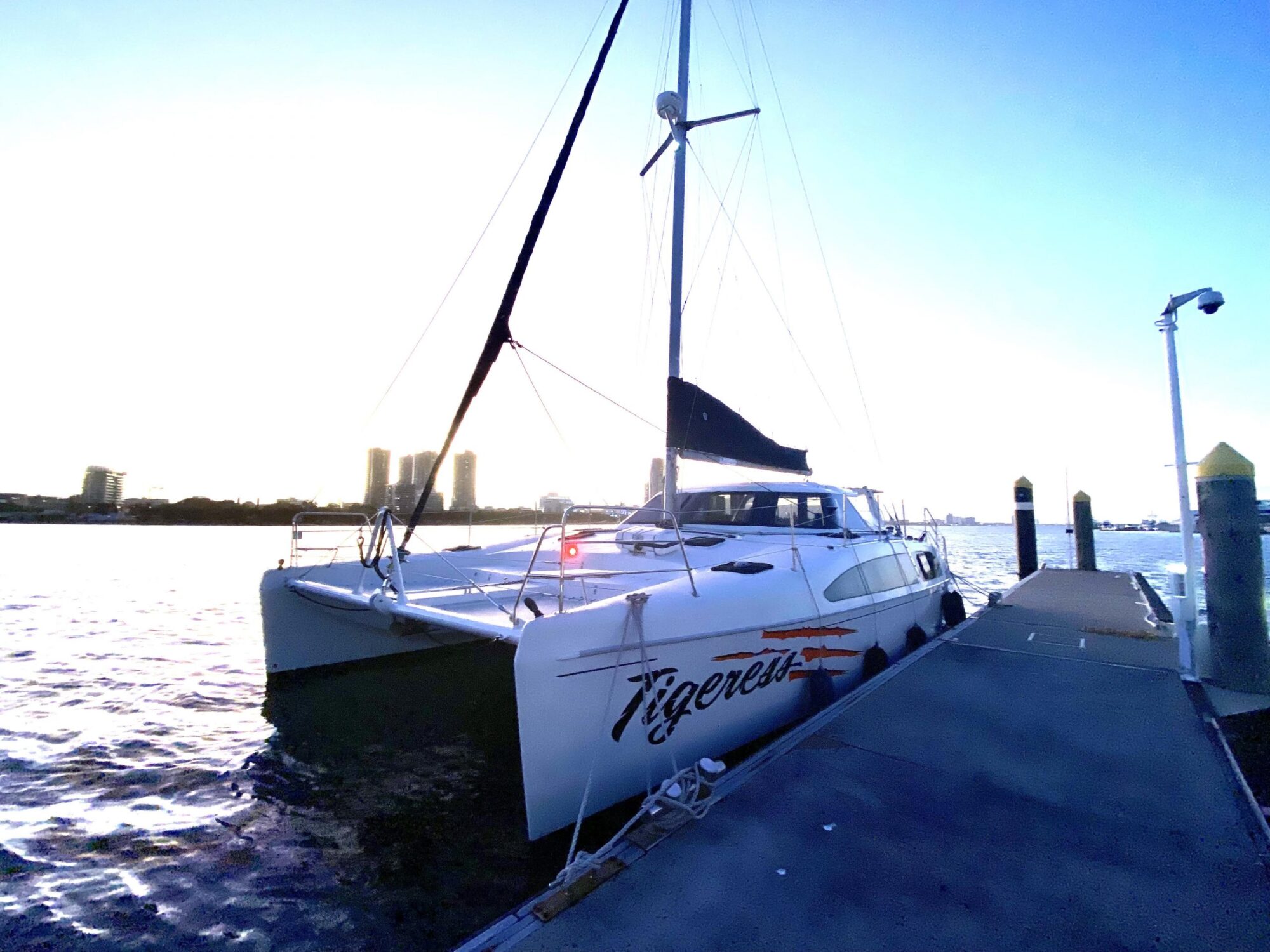 A catamaran named "Tigeress" is docked at a pier during sunset. The sailboat has white hulls and a black sail. The water is calm, and tall buildings are visible in the distance under a clear blue sky.