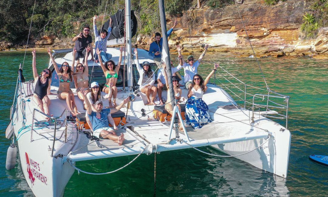 A group of people is smiling and waving while seated and standing on a large white catamaran. The boat is anchored in clear, green waters near a rocky, tree-lined shore. Everyone appears to be enjoying a sunny day out on the water, with laughter ringing through the air.