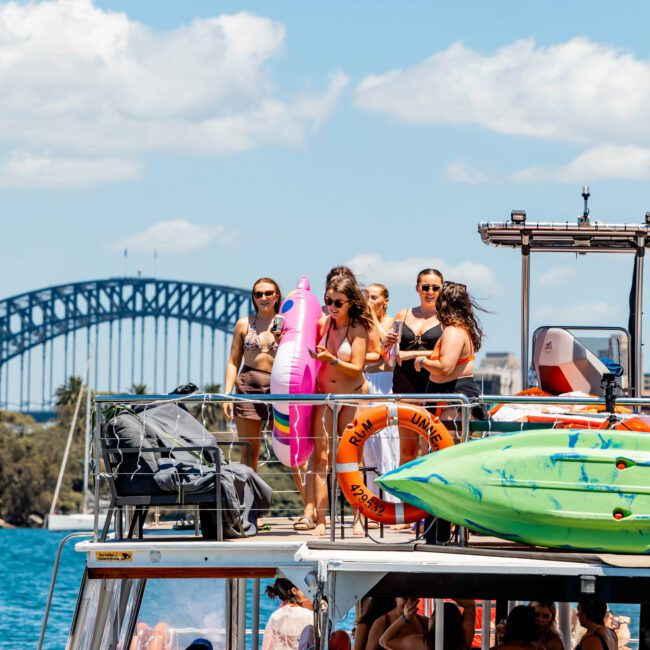 A group of people enjoy a sunny day on a luxury yacht adorned with inflatable toys and colorful paddleboards, courtesy of The Yacht Social Club Event Boat Charters. They are gathered on the upper deck, with a woman holding a pink flamingo float. The background features a large bridge and clear blue skies.