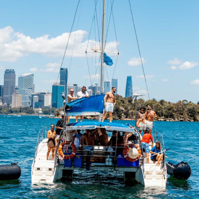 A large sailboat filled with people enjoying a sunny day on the water. The boat, part of The Yacht Social Club, is anchored with a city skyline and blue sky in the background. Some people are on the upper deck, while others lounge on the lower deck and swim near the boat.