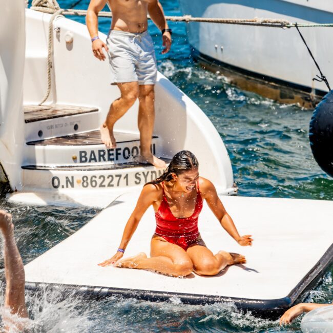 A woman in a red swimsuit sits on a floating platform in the water, smiling. A man in white shorts stands on the boat steps nearby with "BAREFOOT" and "O.N 862271 SYD" on the boat. Another person splashes in the water. A yacht is visible in the background, likely from The Yacht Social Club Sydney Boat Hire.