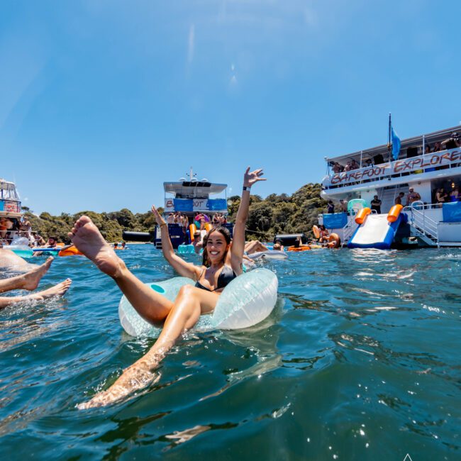 A person lounges on an inflatable tube in the water, raising their arms and smiling. Other people and boats are visible in the background, with one boat displaying the text "Explore." The scene is set on a sunny day, perfect for a day with The Yacht Social Club Sydney Boat Hire.