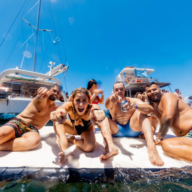 A group of five people sits on the edge of a boat, smiling and reaching towards the camera. The surrounding water is clear, and other boats with additional people are visible in the background. It’s a sunny day with a bright blue sky. Text in the corner reads, "The Yacht Social Club: Boat Parties Sydney.