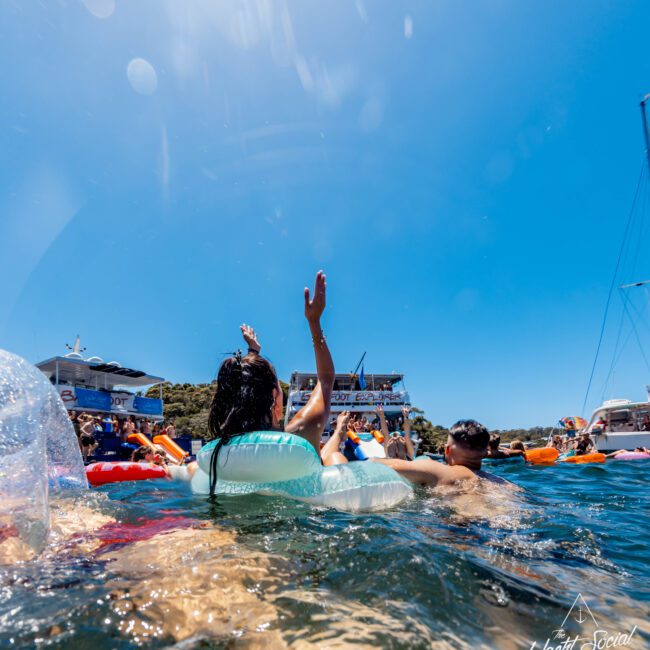 People enjoying a sunny day on the water, some on inflatable tubes and floats, with boats anchored nearby. One person raises their hand in celebration. Clear blue sky and serene water set a festive scene, typical of The Yacht Social Club Event Boat Charters.