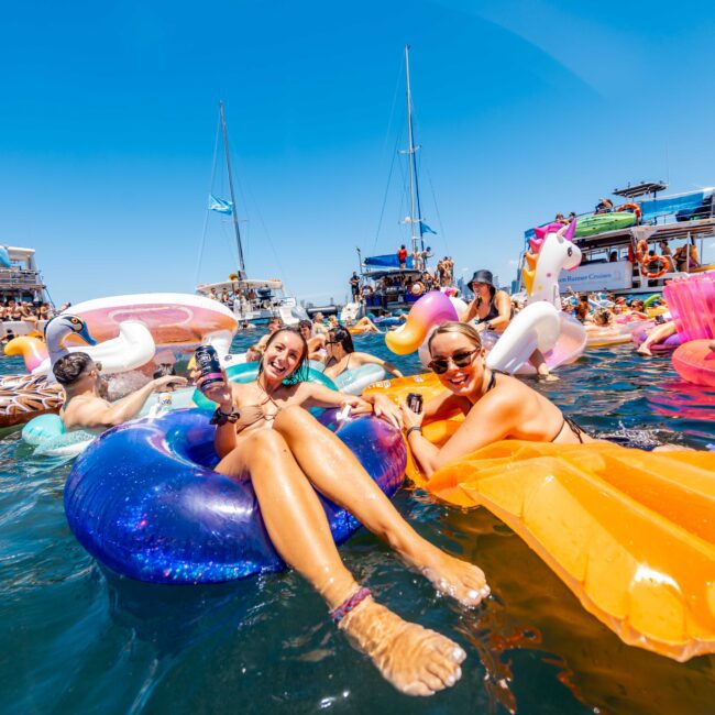 People are lounging on inflatable floats in a sunny, clear blue water setting with yachts in the background. Some inflatables are shaped like animals like a unicorn and a flamingo. Everyone seems to be enjoying the weather and the festivities at The Yacht Social Club's luxury yacht rentals event in Sydney.