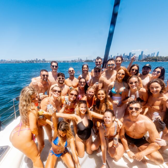 A large group of people in swimsuits poses together on a luxury yacht under a clear blue sky. The water is visible in the background, along with city skyscrapers on the horizon. Everyone appears to be smiling and enjoying the sunny day, epitomizing the vibes of Boat Parties Sydney The Yacht Social Club.