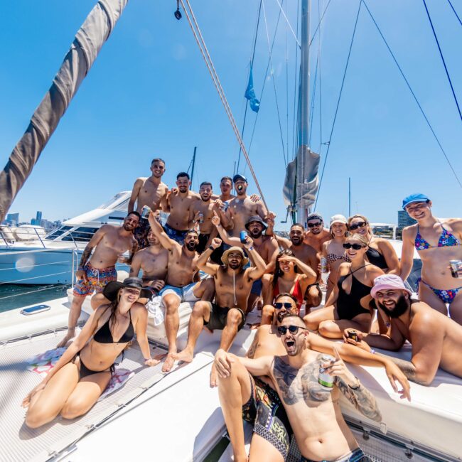 A large group of people in swimwear posing and smiling on the deck of a sailboat on a sunny day. The boat's mast and blue sky are visible in the background. The group appears to be having fun, enjoying their time on the water with The Yacht Social Club Event Boat Charters.