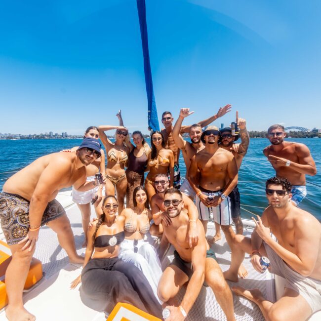 A group of people in swimwear are smiling, posing, and making playful gestures on a luxury yacht in sunny weather. The ocean and a city skyline can be seen in the background. Some are standing, while others are seated on the deck near orange cushions, enjoying the vibes at The Yacht Social Club.