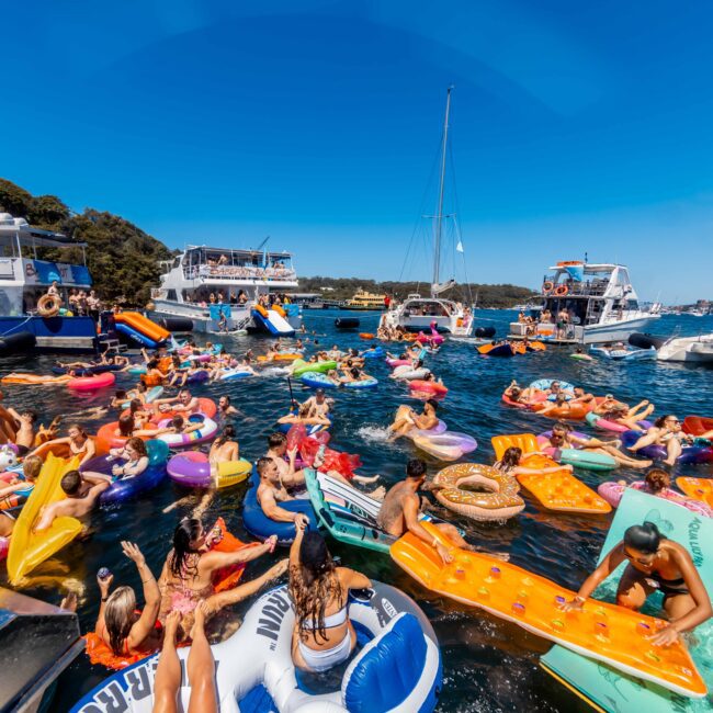 A lively boat party hosted by The Yacht Social Club with people on colorful inflatables in the water between several docked boats. Attendees are enjoying the sunshine and clear blue sky, creating a festive atmosphere. The scene is vibrant with a variety of floaties like unicorns and pizzas.