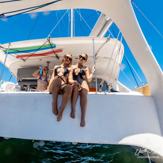 Three people are enjoying their time on a sailboat from Sydney Harbour Boat Hire The Yacht Social Club. Two women in black swimsuits and sunglasses sit on the boat's edge, smiling at the camera, while a third person in a bikini stands in the background under bright, sunny weather with clear blue skies above.