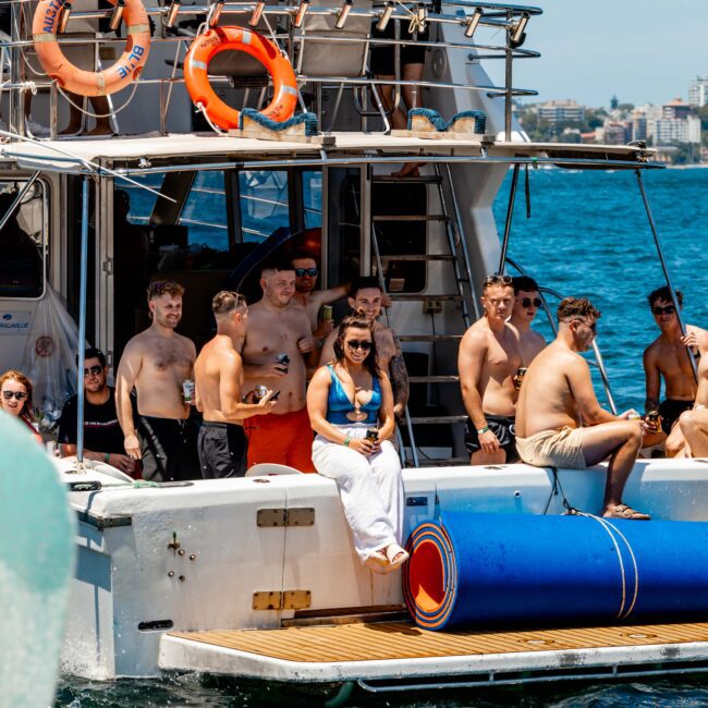 A group of people gathered on a yacht and the water beside it, enjoying a sunny day. Some are sitting on the edge of the yacht, while others are in or near the water. Pool floats and a buoy are also visible. A banner reads "Boat Parties Sydney The Yacht Social Club.