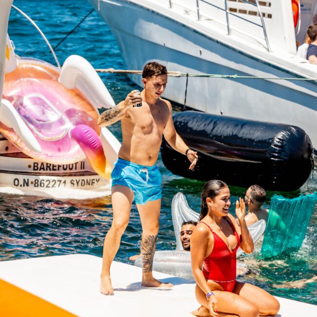 People enjoying a sunny day on the water with inflatables and boats. One man in blue shorts stands on a floating platform, a woman in a red swimsuit sits next to him, while others swim nearby. Boats and a large inflatable are visible in the background, showcasing The Yacht Social Club Sydney Boat Hire experience.