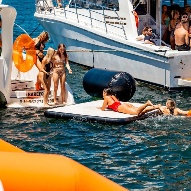 A group of people enjoy a sunny day on the water with yachts in the background. Some are on inflatable floats and others are on the yachts. The scene is lively with people swimming, sunbathing, and socializing, reminiscent of a gathering hosted by The Yacht Social Club Sydney Boat Hire.