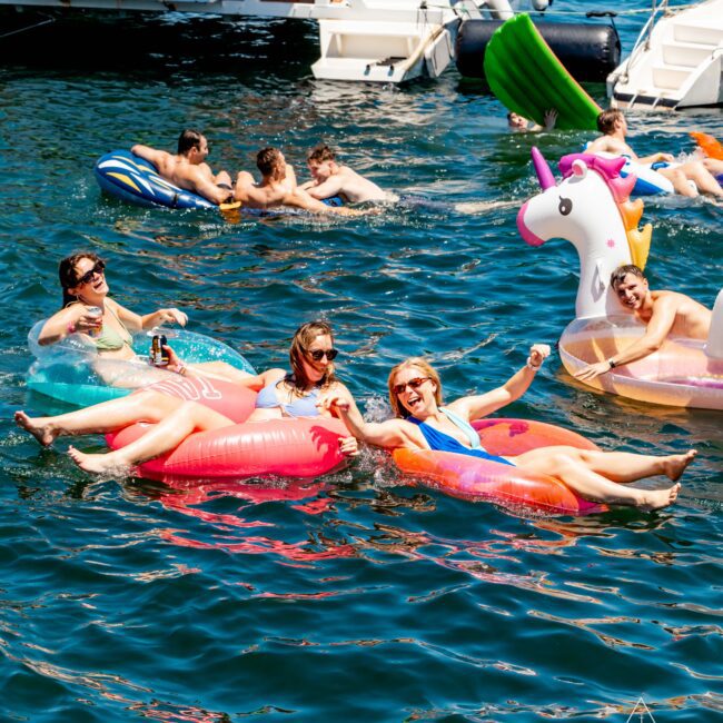 A group of people enjoys a sunny day on a lake, floating on inflatable tubes, including a pink flamingo and a unicorn. In the background, more people swim and lounge on floats near a white boat labeled "Rum Runner Cruises" from The Yacht Social Club Sydney Boat Hire. Smiles and relaxation abound.