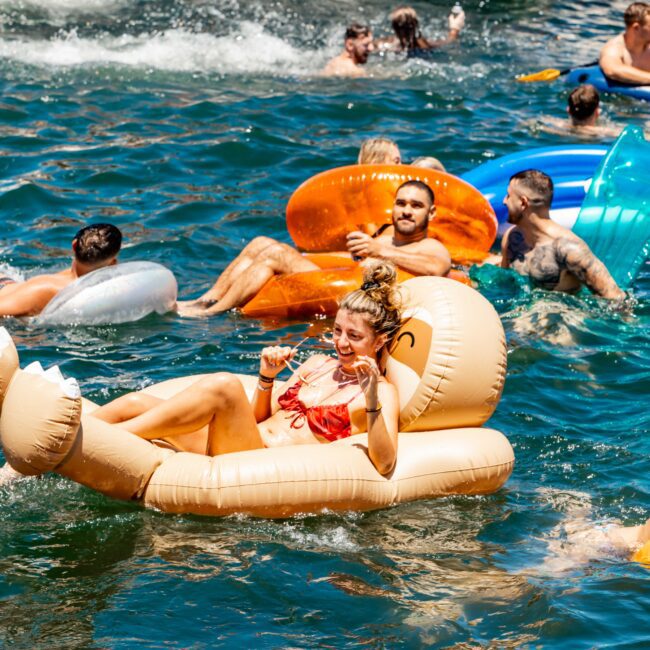 A group of people enjoys a sunny day on the water, floating on inflatable rafts and swimming near a boat. One woman in a red swimsuit relaxes on an inflatable llama raft, smiling and posing for the camera. The Yacht Social Club event offers an unforgettable experience, with the boat and water visible in the background.