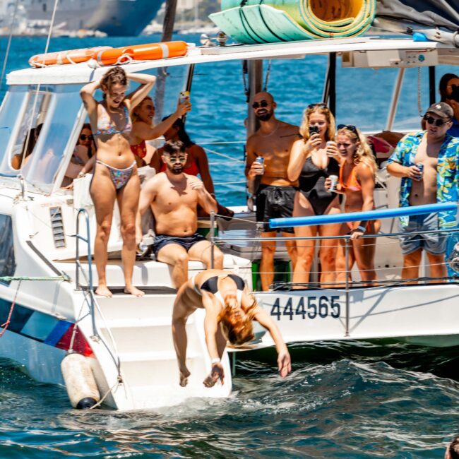 A lively group of people on a boat enjoying a sunny day. A woman in a bikini is diving into the water while others are standing and smiling. The city skyline and other boats are visible in the background. Text in the image: "The Yacht Social Club Event Boat Charters.