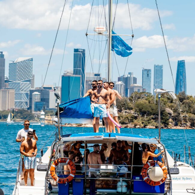 A group of people is enjoying a sunny day on a yacht from The Yacht Social Club Sydney Boat Hire. Some stand on the deck, while others are inside or sitting near the edge. The yacht glides across the water, with a city skyline and other boats in the background. Blue skies and calm waters surround them.