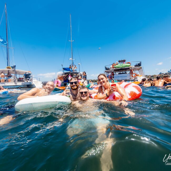 A group of people floats in the water on inflatable rings, smiling and posing for the camera. Behind them are several boats and more people enjoying the water. The sky is clear and blue, indicating a sunny day. "The Yacht Social Club Event Boat Charters" are visible in the bottom right corner.