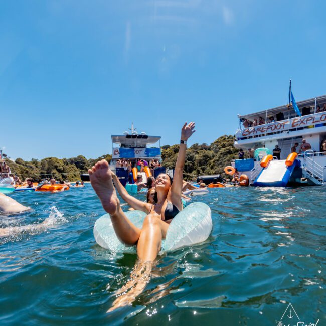 A person floats on an inflatable tube in clear blue water, laughing with arms raised. In the background, people enjoy water activities near a luxury yacht. Bright, sunny day with a clear sky. Text on the image reads "The Yacht Social Club".