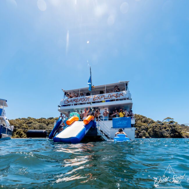 The "Barefoot Explorer" from Luxury Yacht Rentals Sydney is anchored near a lush island. With a slide extending from its upper deck into clear blue water, several guests enjoy the sunny day on the boat and in the water. Another boat, possibly from The Yacht Social Club Sydney Boat Hire, is visible in the distance.