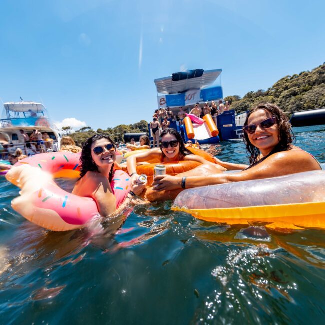 Three people are floating in the water on inflatables, holding drinks and smiling at the camera. Others are in the background, enjoying a sunny day on the water. The scene is lively, with boats and people in swimsuits. The Yacht Social Club Sydney Boat Hire logo is visible in the bottom right.