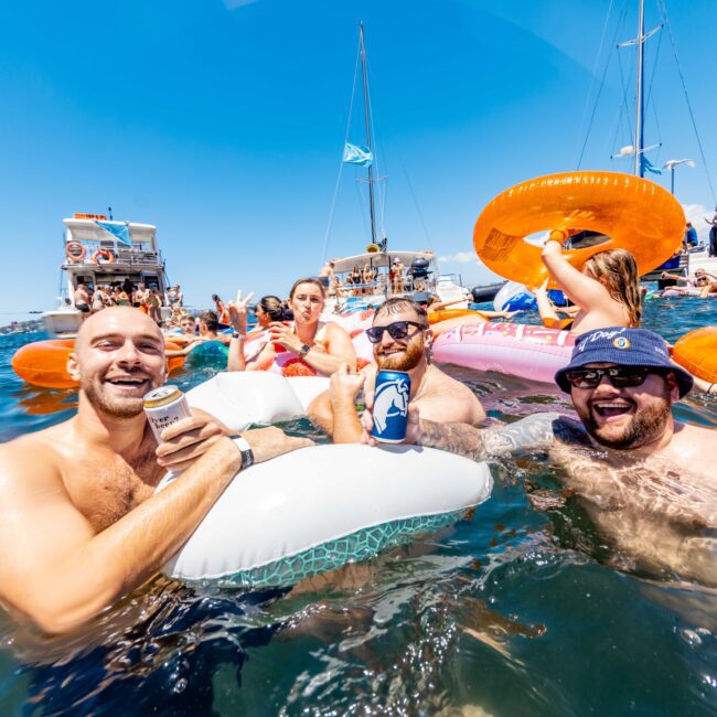 A group of people enjoying a sunny day on the water, with several individuals in the foreground floating on inflatable tubes and holding drinks. Boats and more people can be seen in the background, along with clear blue skies above—a perfect scene from The Yacht Social Club Event Boat Charters.