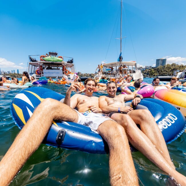 Two people relax on a large pool float among a lively crowd at a yacht party. The water is filled with colorful inflatables, and boats in the background contribute to the festive atmosphere under a bright blue sky. Logo in the corner reads "The Yacht Social Club Sydney Boat Hire.