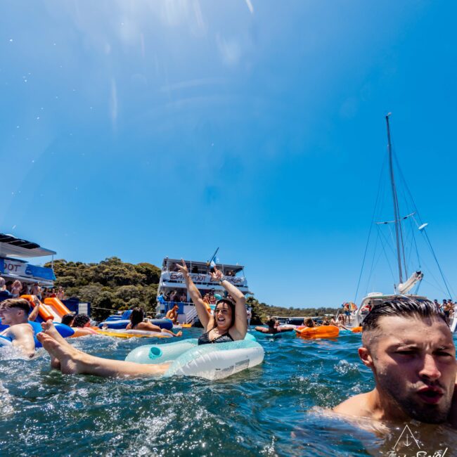 People enjoying a sunny day on the water, with some floating on inflatable devices. A woman joyfully raises her arms on an inflatable, while others are nearby in the water. In the background, boats and more people are visible, exemplifying a delightful Boat Parties Sydney The Yacht Social Club atmosphere. The sky is clear and blue.