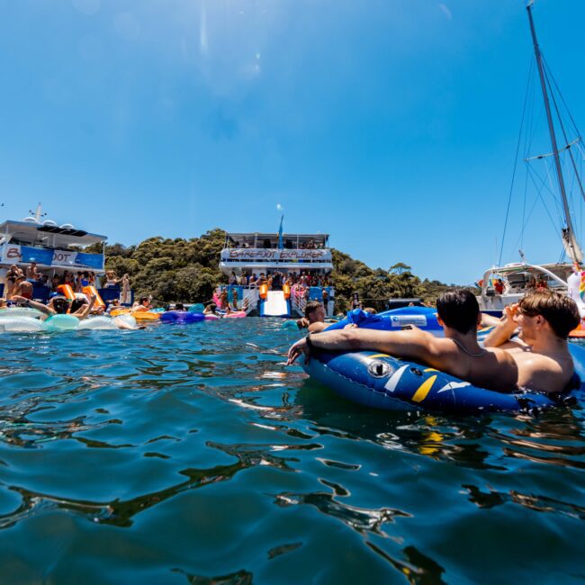 A lively boat party scene on a sunny day shows people relaxing on inflatable tubes in the water, with several yachts and a floating stage in the background. The clear blue sky and calm waters add to the festive atmosphere. The Yacht Social Club Sydney Boat Hire logo is visible.