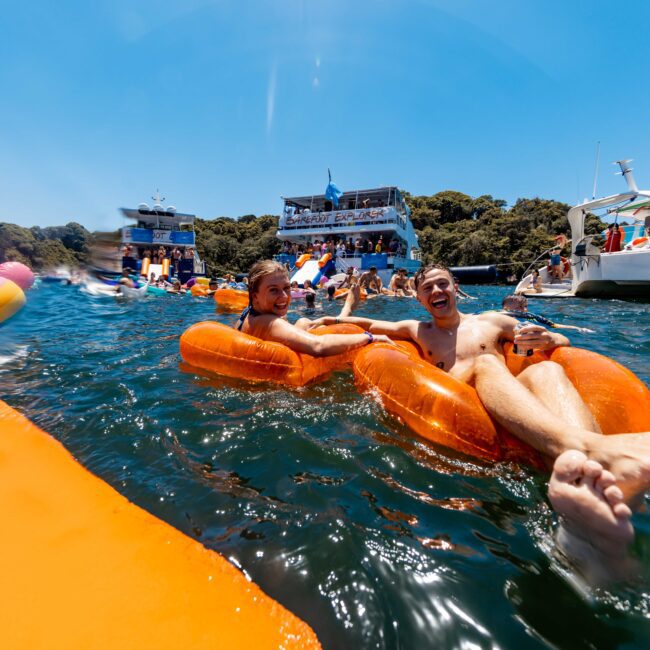 A group enjoys a sunny day on the water, floating on inflatable loungers near boats. Two men on orange loungers smile and hold drinks. In the background, people socialize on a boat with a banner. Text: "Yacht Social Club." Experience unforgettable moments with The Yacht Social Club Event Boat Charters.