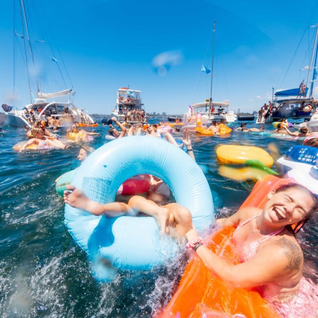 A large group of people are enjoying a sunny day on the water, surrounded by boats and colorful inflatables. Many are swimming, floating, and laughing. The atmosphere is lively and festive, with clear blue skies and sparkling water in the background, perfect for a Luxury Yacht Rentals Sydney experience.