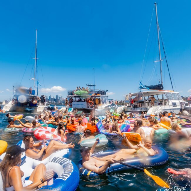 A lively scene of people enjoying a sunny day on inflatable pool floats and boats on the water. The crowd appears to be having fun, with some people holding drinks. In the background, a city skyline is visible under a clear blue sky. This vibrant atmosphere is reminiscent of the Yacht Social Club Sydney boat parties.