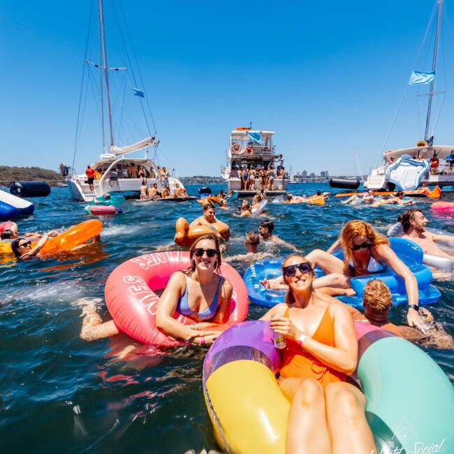 People enjoying a sunny day floating on colorful inflatables in the water. Several yachts and boats, courtesy of Luxury Yacht Rentals Sydney, are anchored close by, with more people socializing on them. Blue skies overhead and a fun, lively atmosphere prevail.