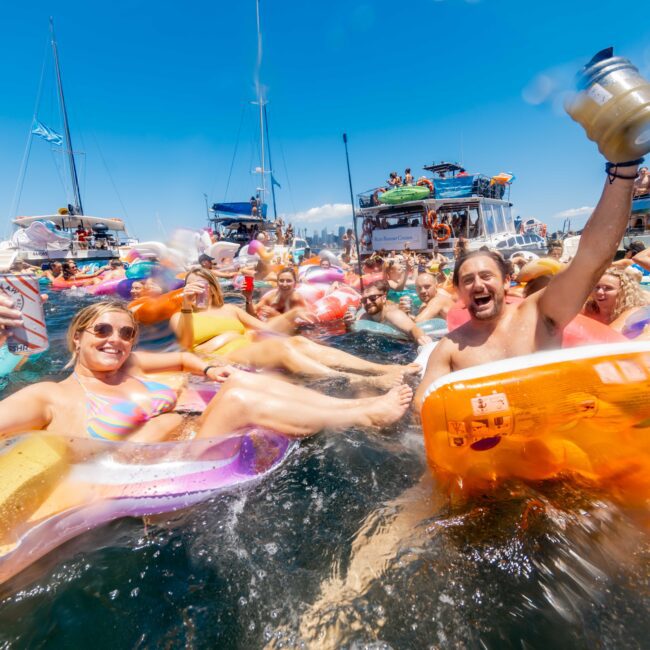 A lively group of people float on inflatable rafts while holding drinks and partying in the water at The Yacht Social Club Sydney Boat Hire. Boats are visible in the background on a sunny day. The image captures energetic expressions and colorful inflatables, creating a festive and vibrant atmosphere.