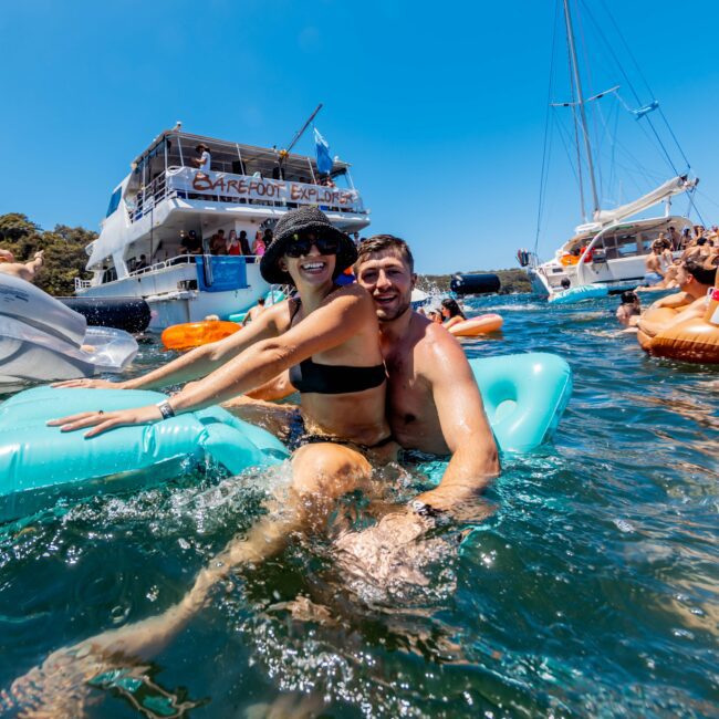 A smiling couple enjoys a sunny day on a turquoise inflatable raft in the water. Behind them are boats and other people relaxing on inflatables. The water is clear, and the sky is bright blue. The logo of "The Yacht Social Club" is visible in the bottom right corner, promoting Sydney Harbour Boat Hire.