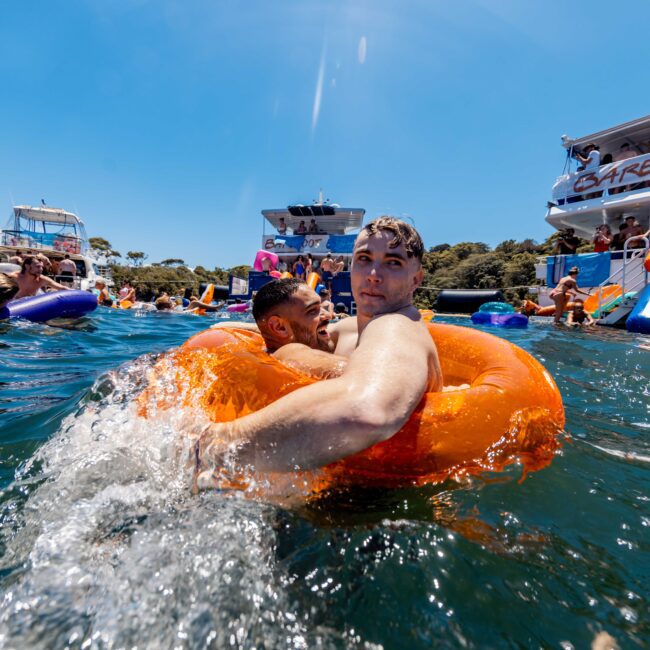 Two people float on an orange inner tube in a crowded, sunny water party scene. Around them, others enjoy various flotation devices near boats. In the background, a large boat with a waterslide and "Barefoot" written on a banner can be seen—a quintessential Boat Parties Sydney The Yacht Social Club moment.