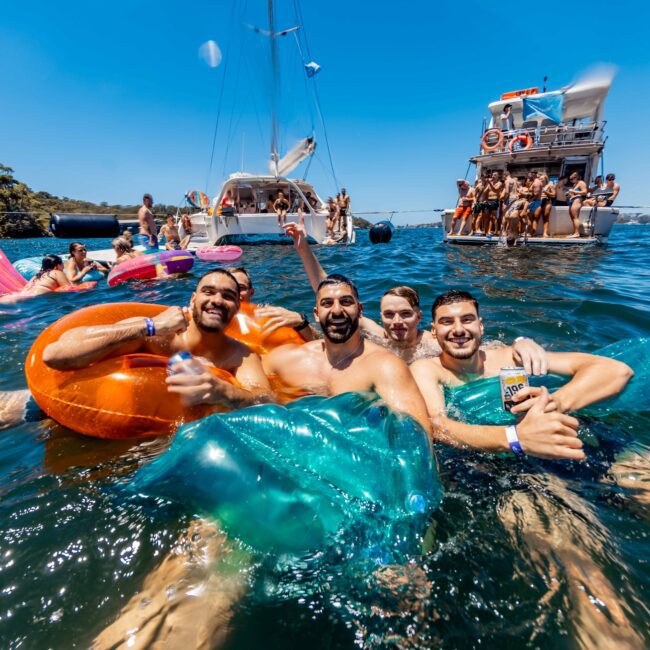 A group of four smiling men floats on inflatable rafts in the water, enjoying a sunny day. Behind them, more people relax on boats and swim, showcasing Boat Parties Sydney The Yacht Social Club. Trees and a clear blue sky form the background. Everyone appears to be having a fun and lively time.