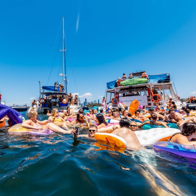 A large group of people enjoying a sunny day on the water at The Yacht Social Club event, floating on inflatable rafts and swimming near anchored boats. The sky is clear and the atmosphere lively, with some people on the boats and others in the water.