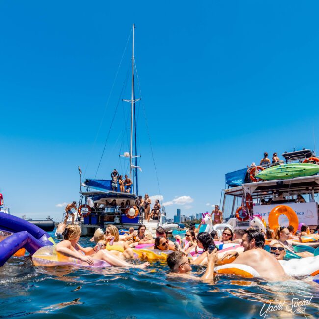 A large group of people enjoying a sunny day on the water with The Yacht Social Club, relaxing on colorful inflatable floats around a boat. The boat is anchored, and some are on deck while others are in the water. The city skyline is visible in the distance under a clear blue sky.