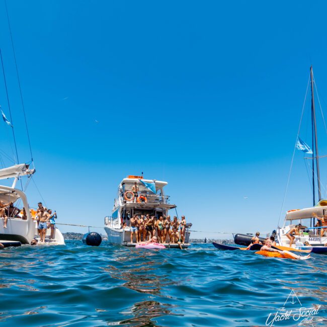 A sunny day on the water with multiple boats anchored close together. People are gathered on the decks and in the water, swimming and enjoying the weather. The sky is clear and blue, creating a picturesque scene of leisure at The Yacht Social Club Event Boat Charters in Sydney Harbour.