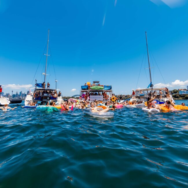 People are swimming and floating in an ocean area surrounded by several anchored yachts and boats under a clear blue sky. The water is calm, with flags waving on some boats. A cityscape is faintly visible in the background, setting the scene for The Yacht Social Club Event Boat Charters.